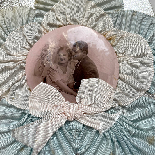 1930s Photo Card showing a romantic couple, surrounded by pleated blue and cream ribbons on a silver textured background.