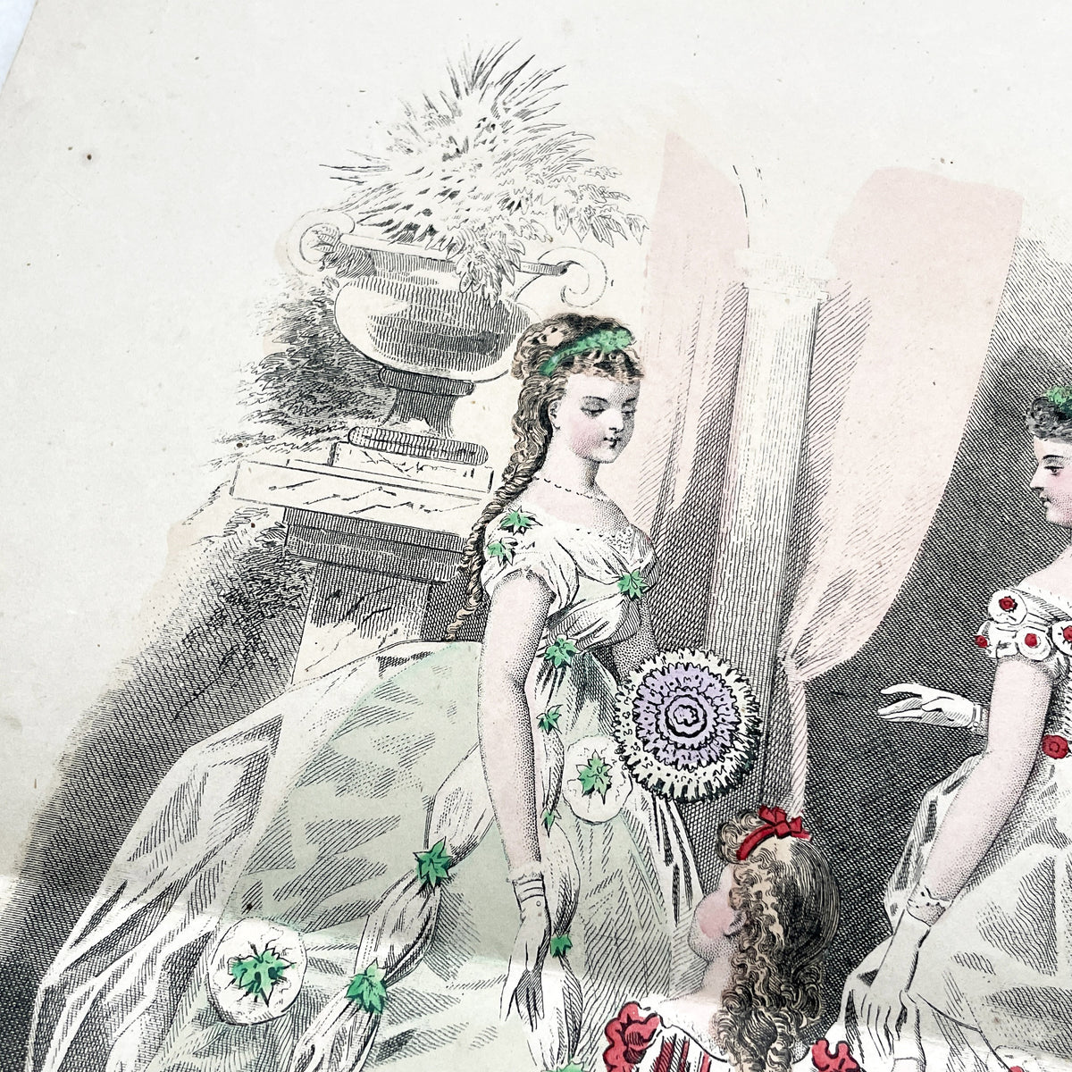 1860s French fashion plate depicting two women in elaborate ball gowns and a child in a red and white striped dress.