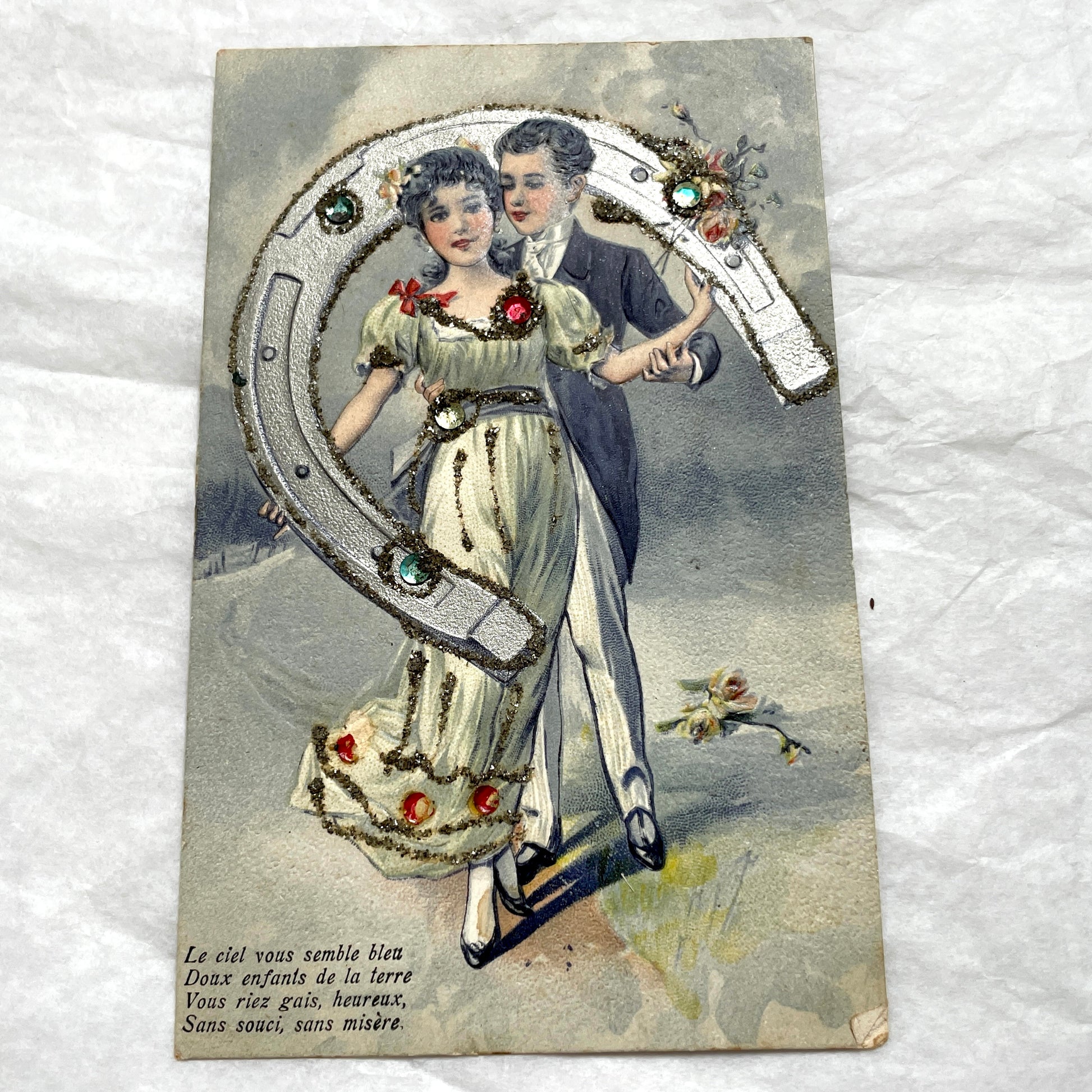 1900s French postcard with a couple holding a glitter horseshoe, adorned with small gems.