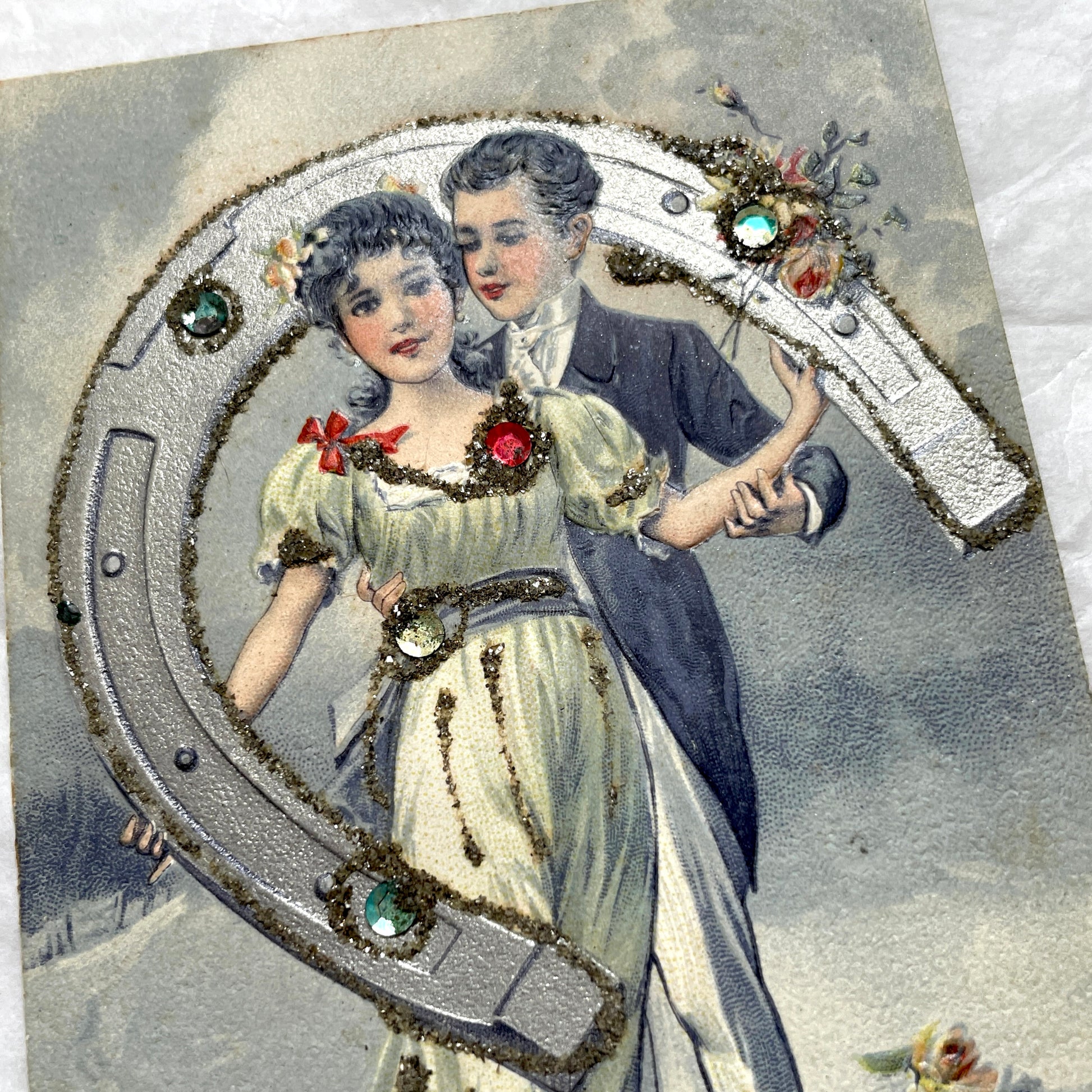 1900s French postcard with a couple holding a glitter horseshoe, adorned with small gems.