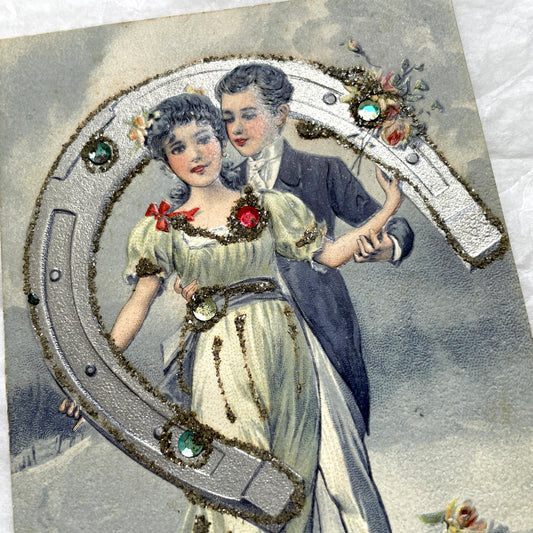 1900s French postcard with a couple holding a glitter horseshoe, adorned with small gems.