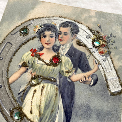 1900s French postcard with a couple holding a glitter horseshoe, adorned with small gems.