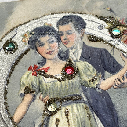 1900s French postcard with a couple holding a glitter horseshoe, adorned with small gems.