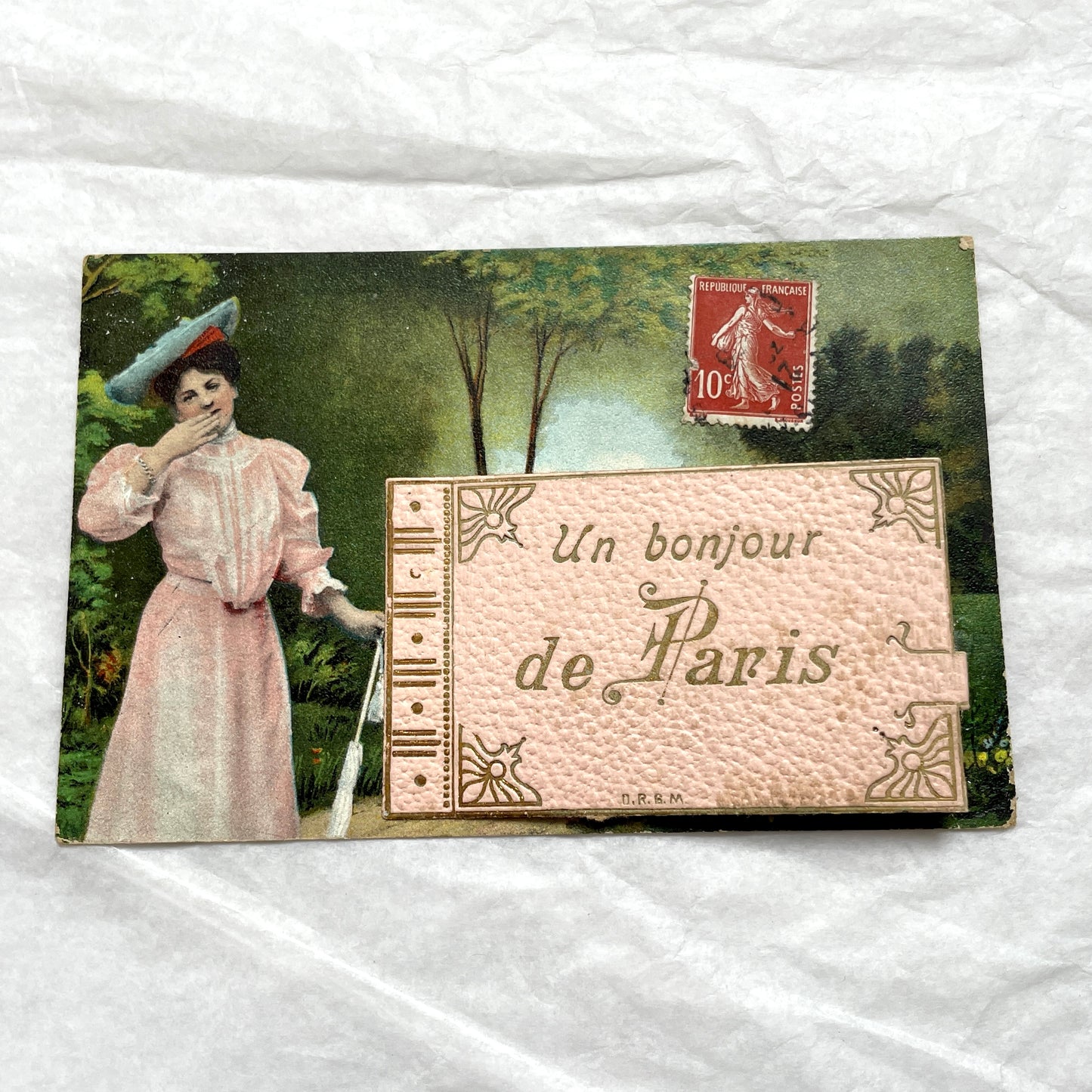 1900s French postcard featuring a Parisian lady in a pink dress, blue hat, with 'Un bonjour de Paris' text.