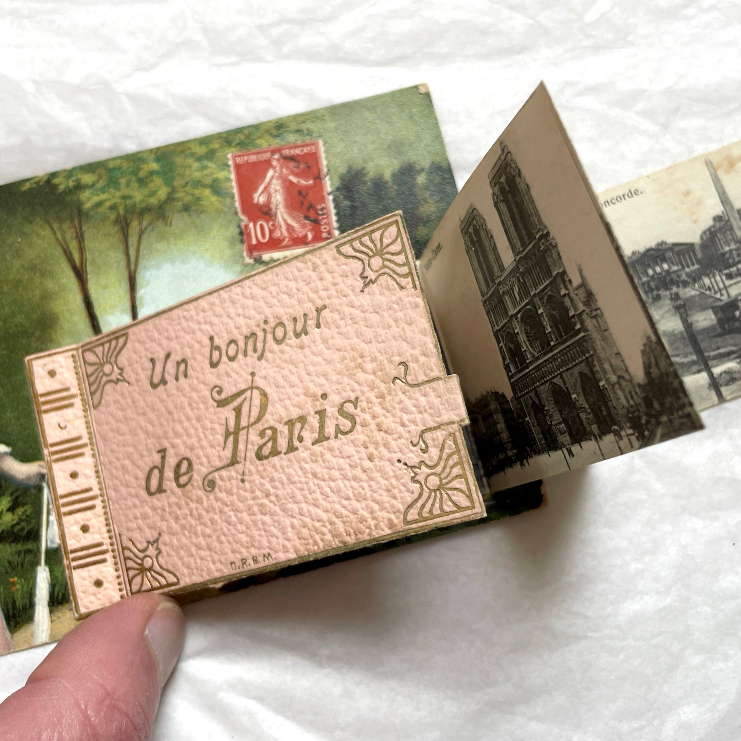 1900s French postcard featuring a Parisian lady in a pink dress, blue hat, with 'Un bonjour de Paris' text.