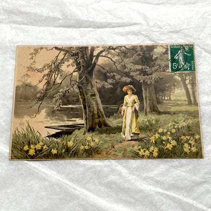 1900s French postcard depicting a woman in a yellow dress walking through a field of daffodils.