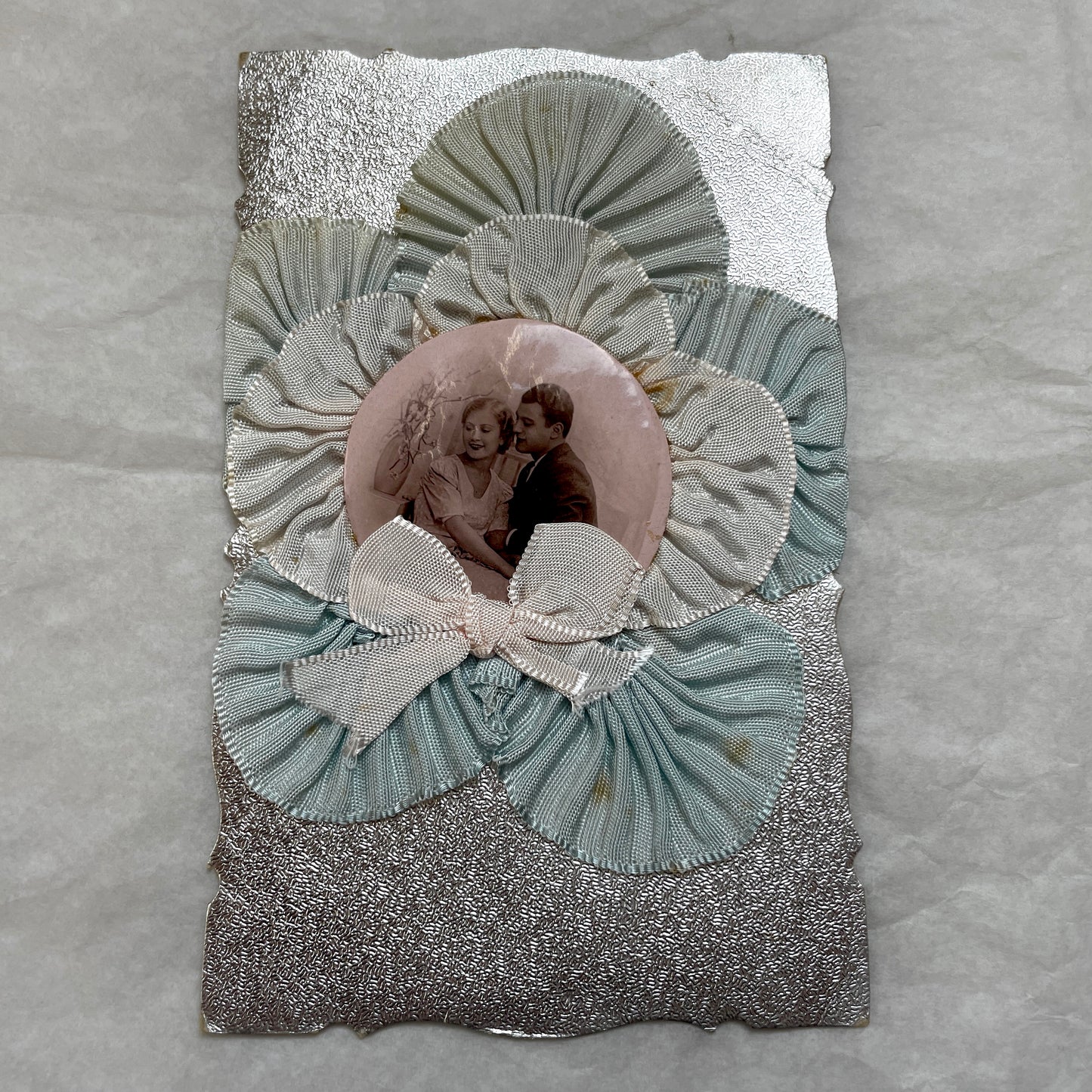 1930s Photo Card showing a romantic couple, surrounded by pleated blue and cream ribbons on a silver textured background.