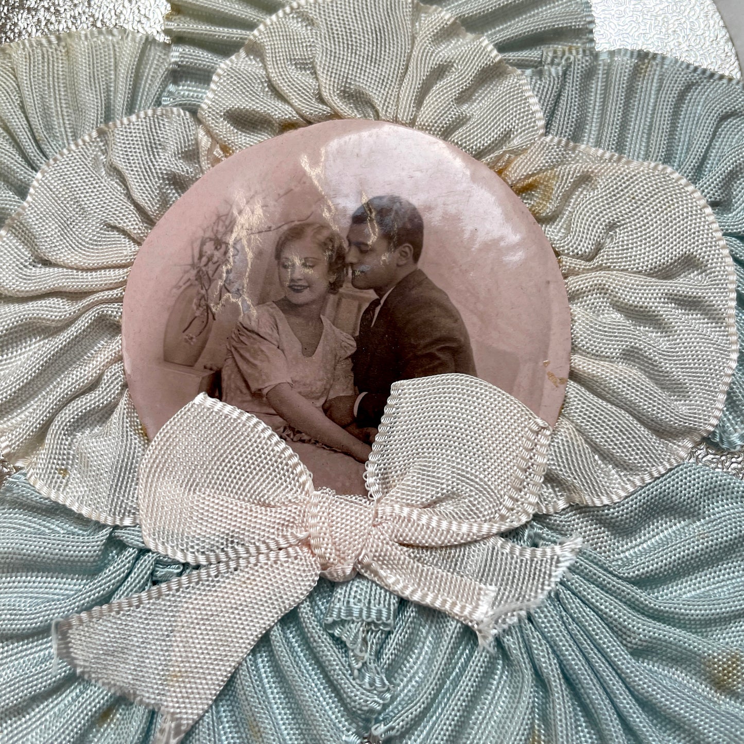 1930s Photo Card showing a romantic couple, surrounded by pleated blue and cream ribbons on a silver textured background.
