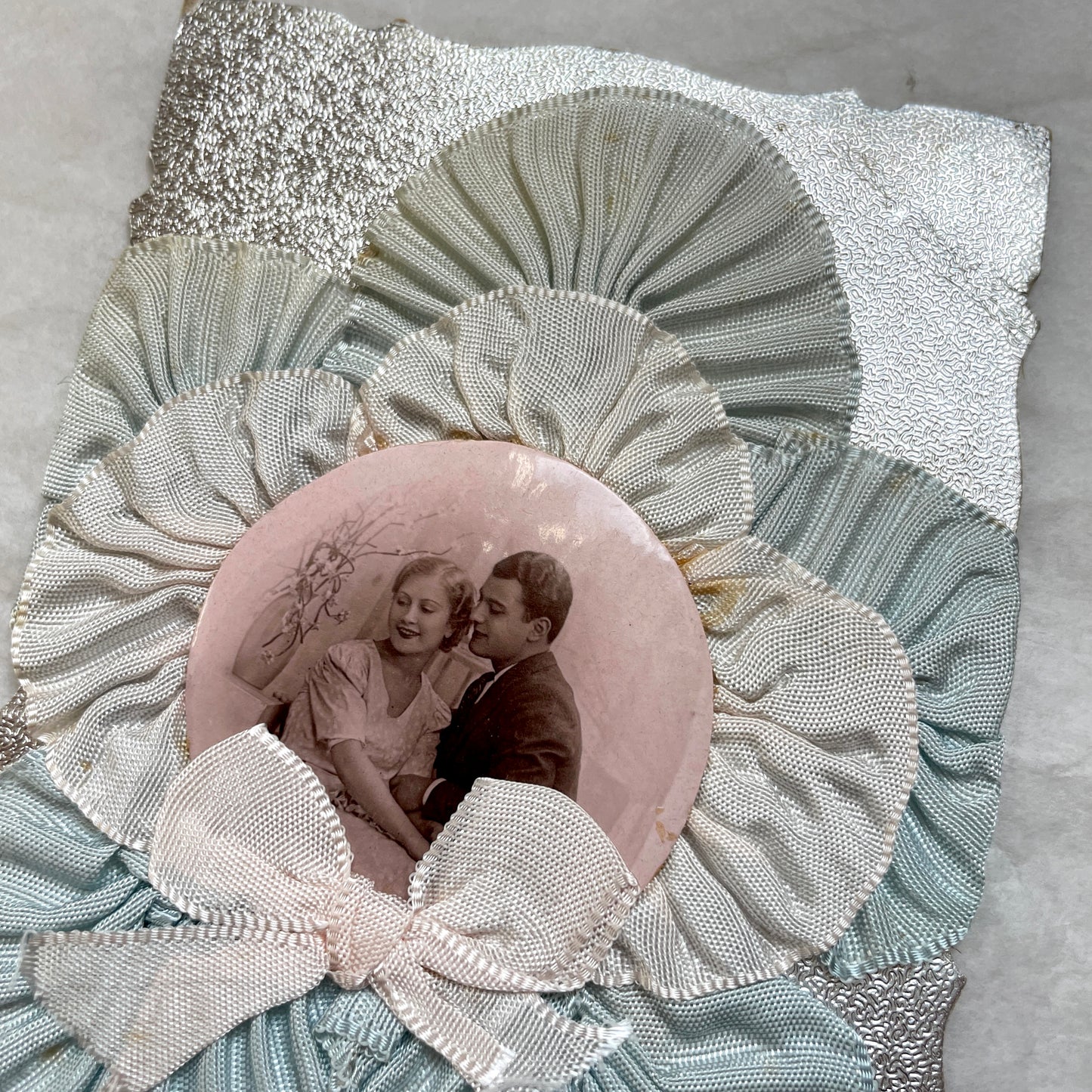 1930s Photo Card showing a romantic couple, surrounded by pleated blue and cream ribbons on a silver textured background.