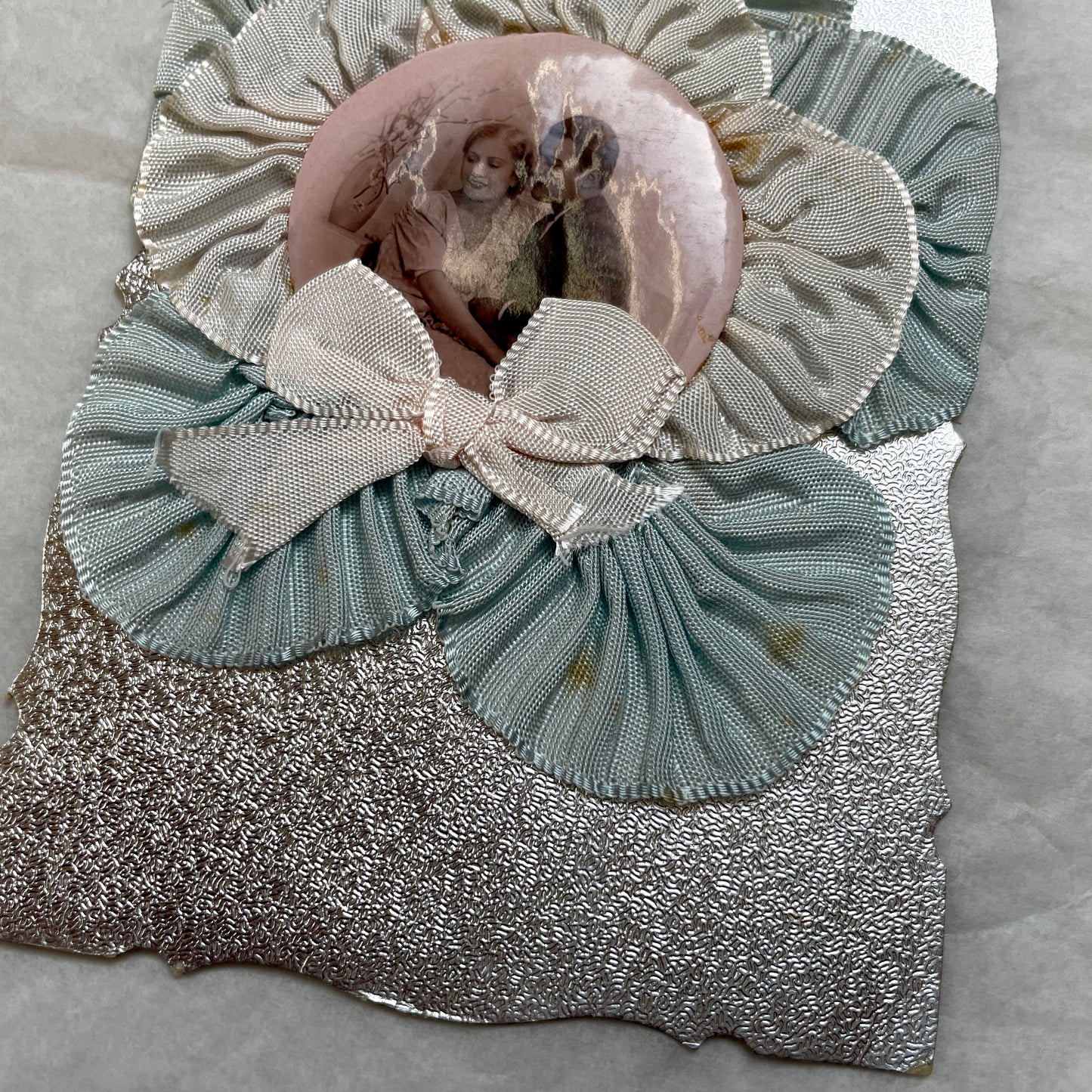 1930s Photo Card showing a romantic couple, surrounded by pleated blue and cream ribbons on a silver textured background.
