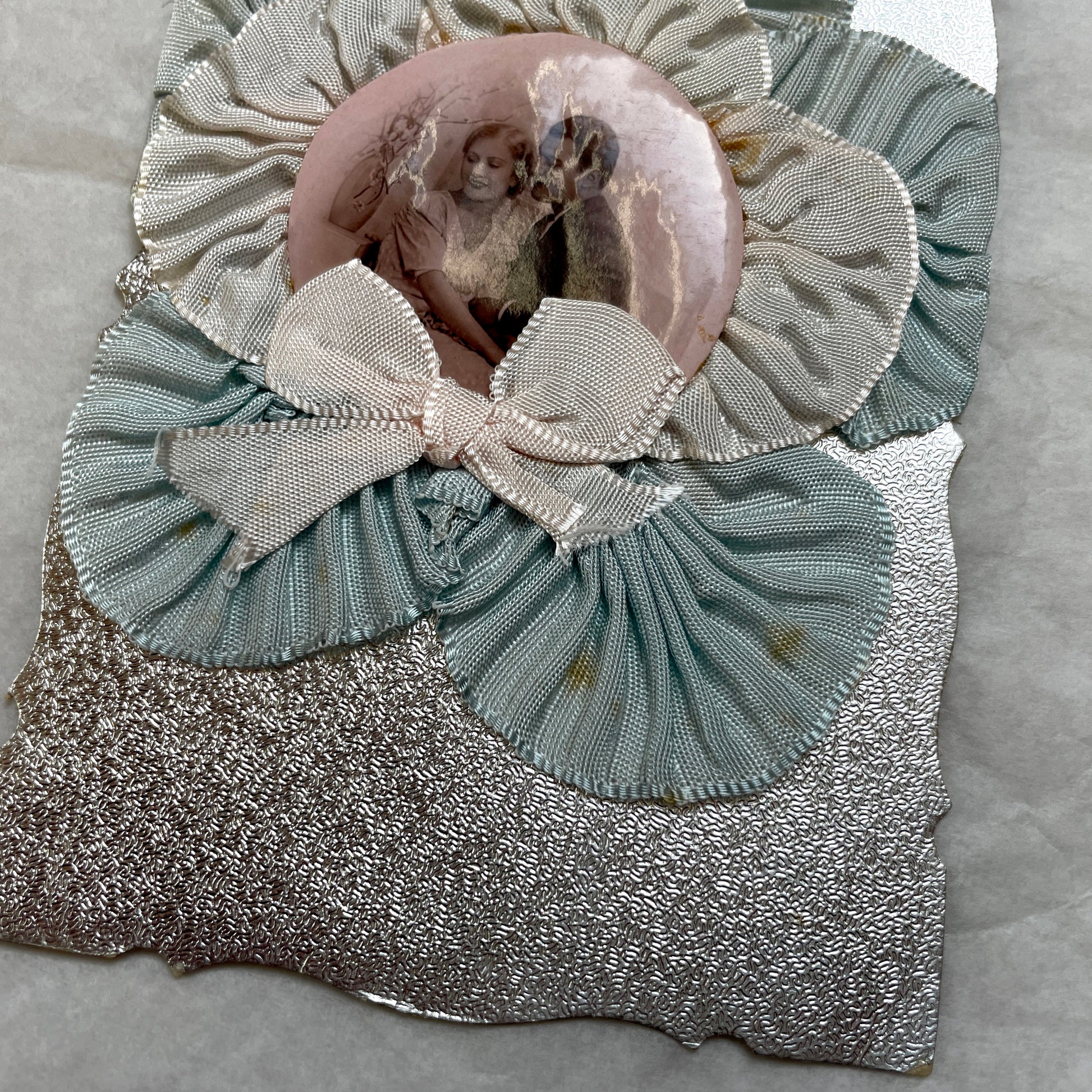 1930s Photo Card showing a romantic couple, surrounded by pleated blue and cream ribbons on a silver textured background.