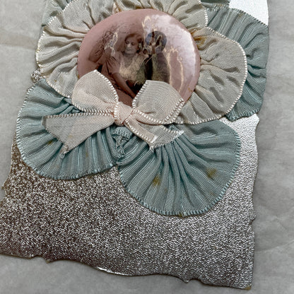 1930s Photo Card showing a romantic couple, surrounded by pleated blue and cream ribbons on a silver textured background.