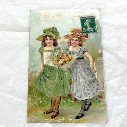 1900s postcard featuring two young girls in garden dresses with hats and a bouquet, adorned with glitter and a French stamp.