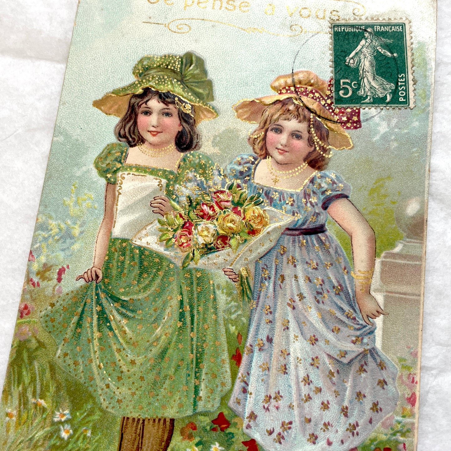 1900s postcard featuring two young girls in garden dresses with hats and a bouquet, adorned with glitter and a French stamp.