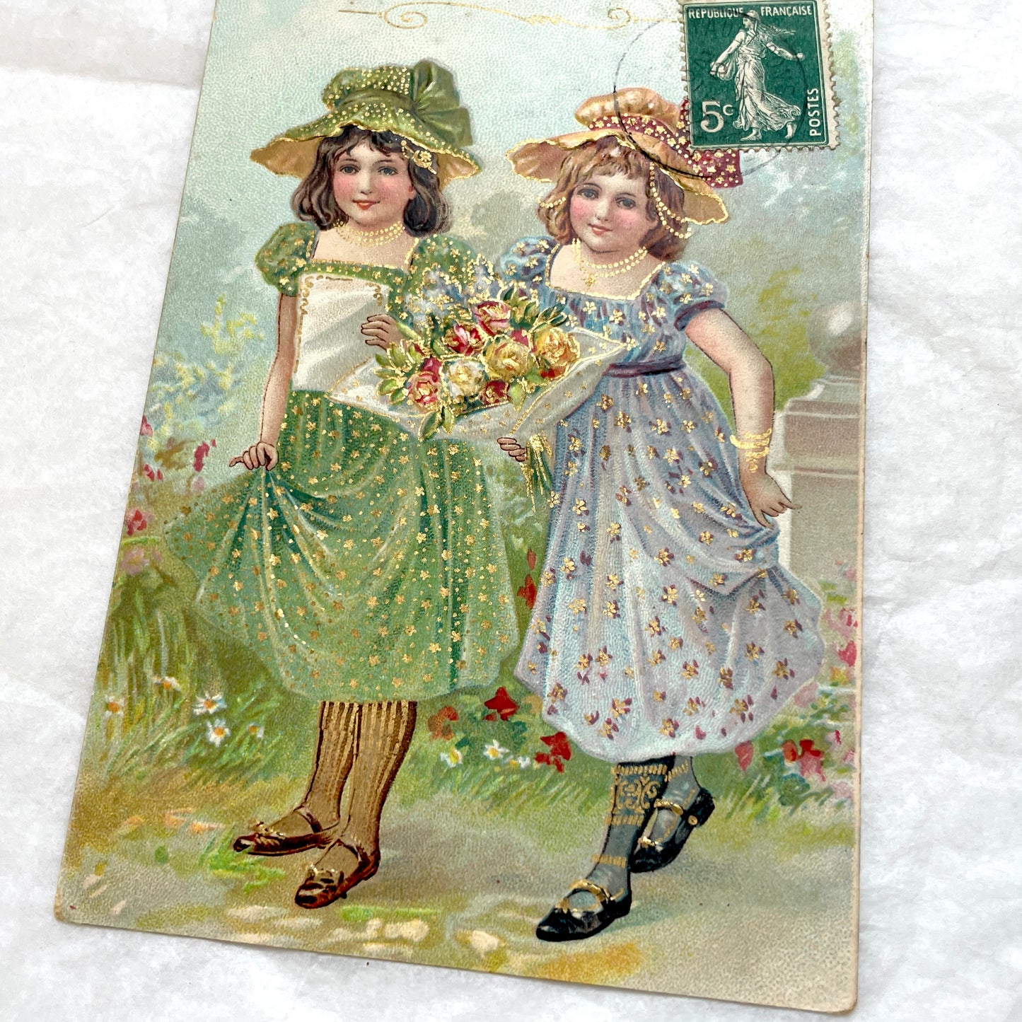1900s postcard featuring two young girls in garden dresses with hats and a bouquet, adorned with glitter and a French stamp.