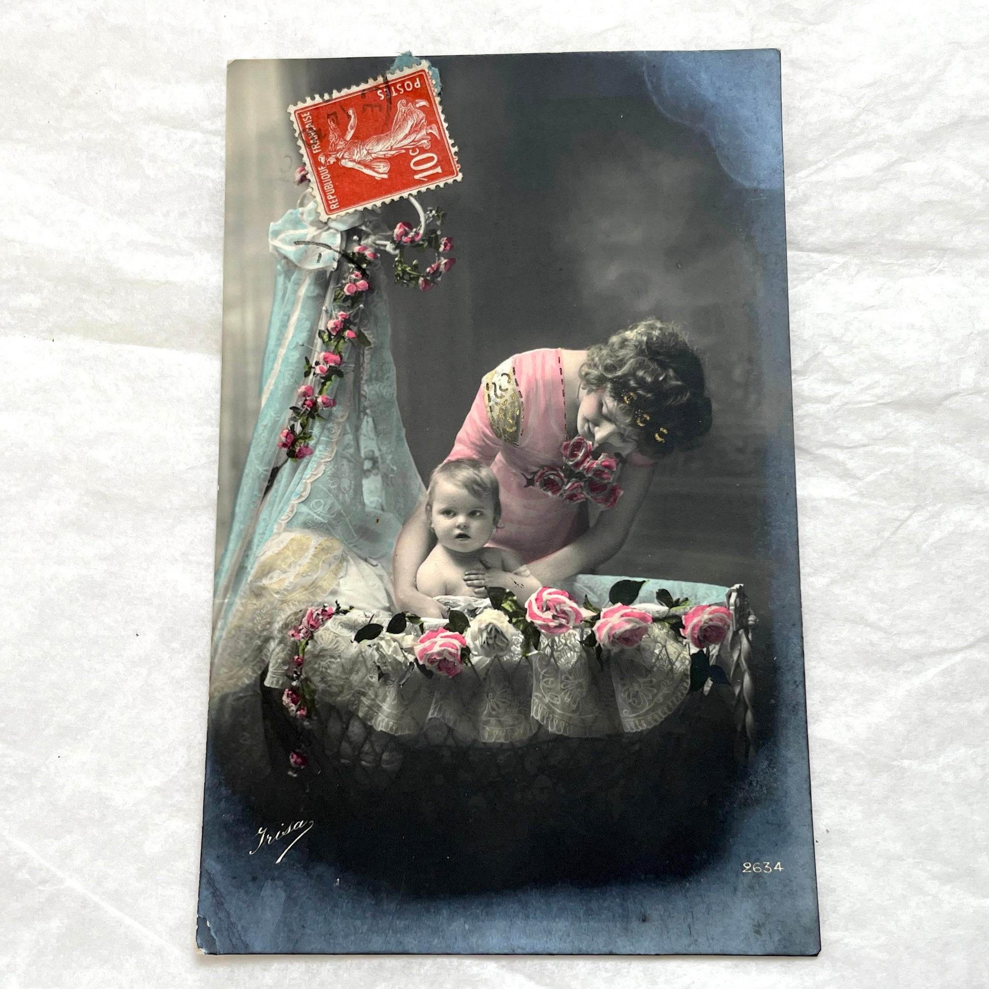 1910s French postcard featuring a hand-colored image of a mother tenderly holding her baby amidst pink roses.