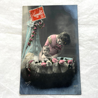 1910s French postcard featuring a hand-colored image of a mother tenderly holding her baby amidst pink roses.
