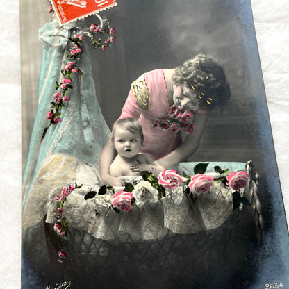1910s French postcard featuring a hand-colored image of a mother tenderly holding her baby amidst pink roses.