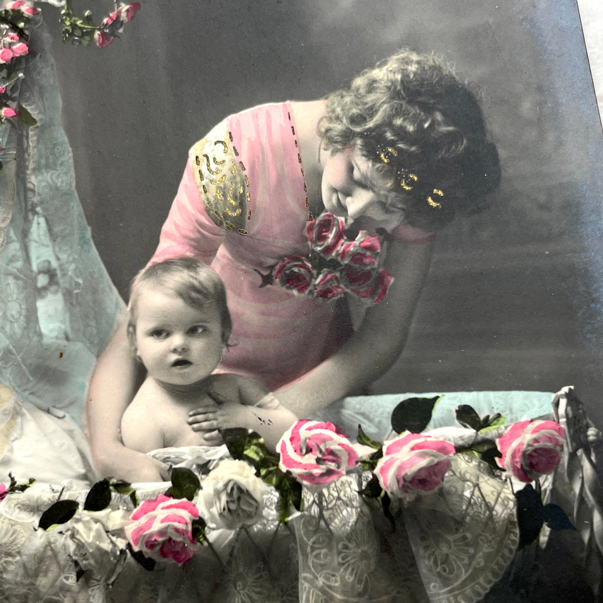 1910s French postcard featuring a hand-colored image of a mother tenderly holding her baby amidst pink roses.
