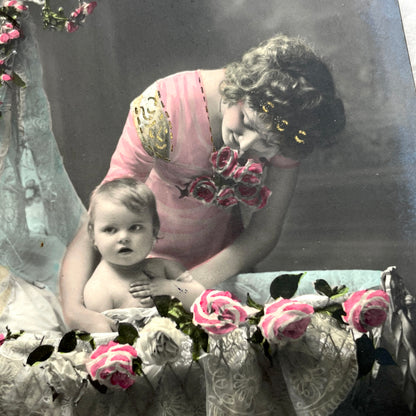 1910s French postcard featuring a hand-colored image of a mother tenderly holding her baby amidst pink roses.
