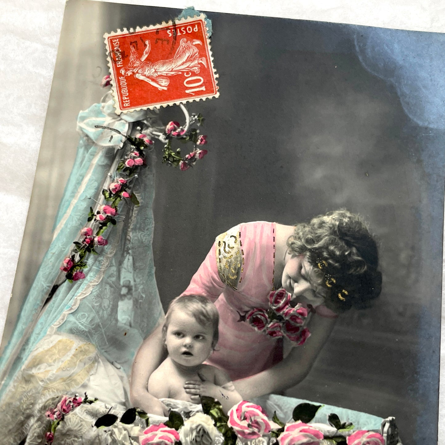 1910s French postcard featuring a hand-colored image of a mother tenderly holding her baby amidst pink roses.